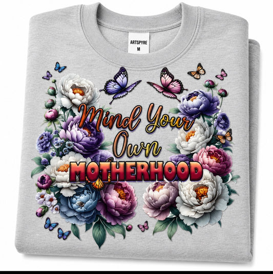 A folded light gray crewneck sweatshirt with a colorful graphic print. The design features the text 'Mind Your Own MOTHERHOOD' surrounded by white, purple, and blue peonies, green leaves, and butterflies.